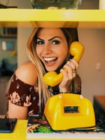 A woman speaks happily into a large yellow telephone.