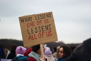 At a protest, a woman holds a sign saying "what lessens one of us lessens all of us".