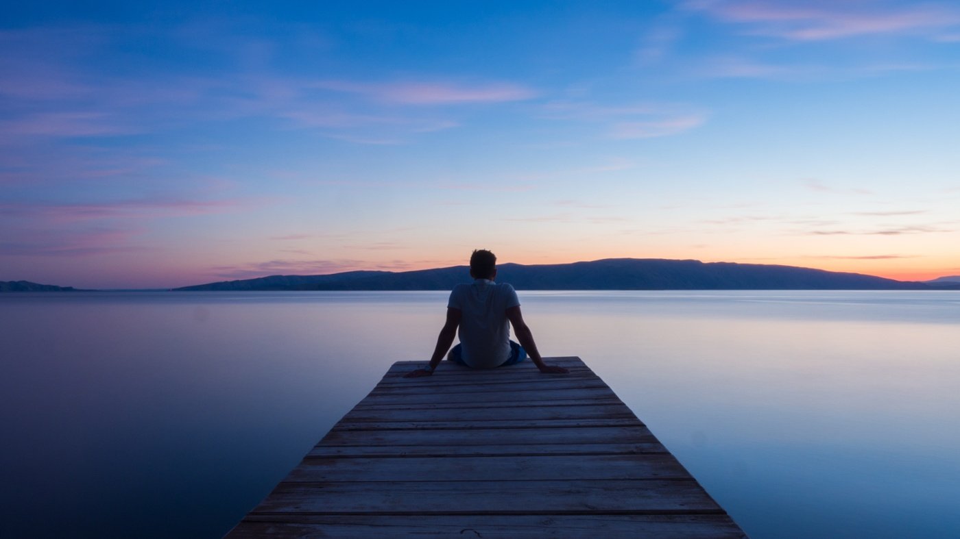 A person sits peacefully at the end of a dock, looking out onto a lake.